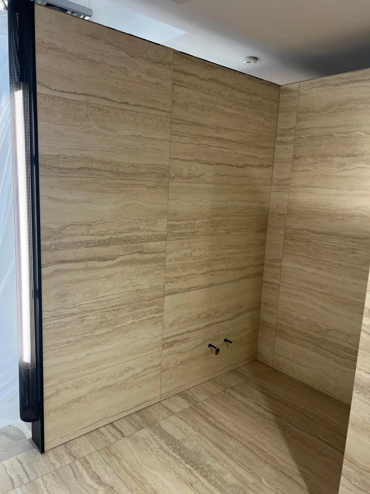 A shower space with beige marble tiles on the walls and floor, two black water pipe fittings visible at the lower part of the back wall, and a partially visible black-framed glass door on the left side.