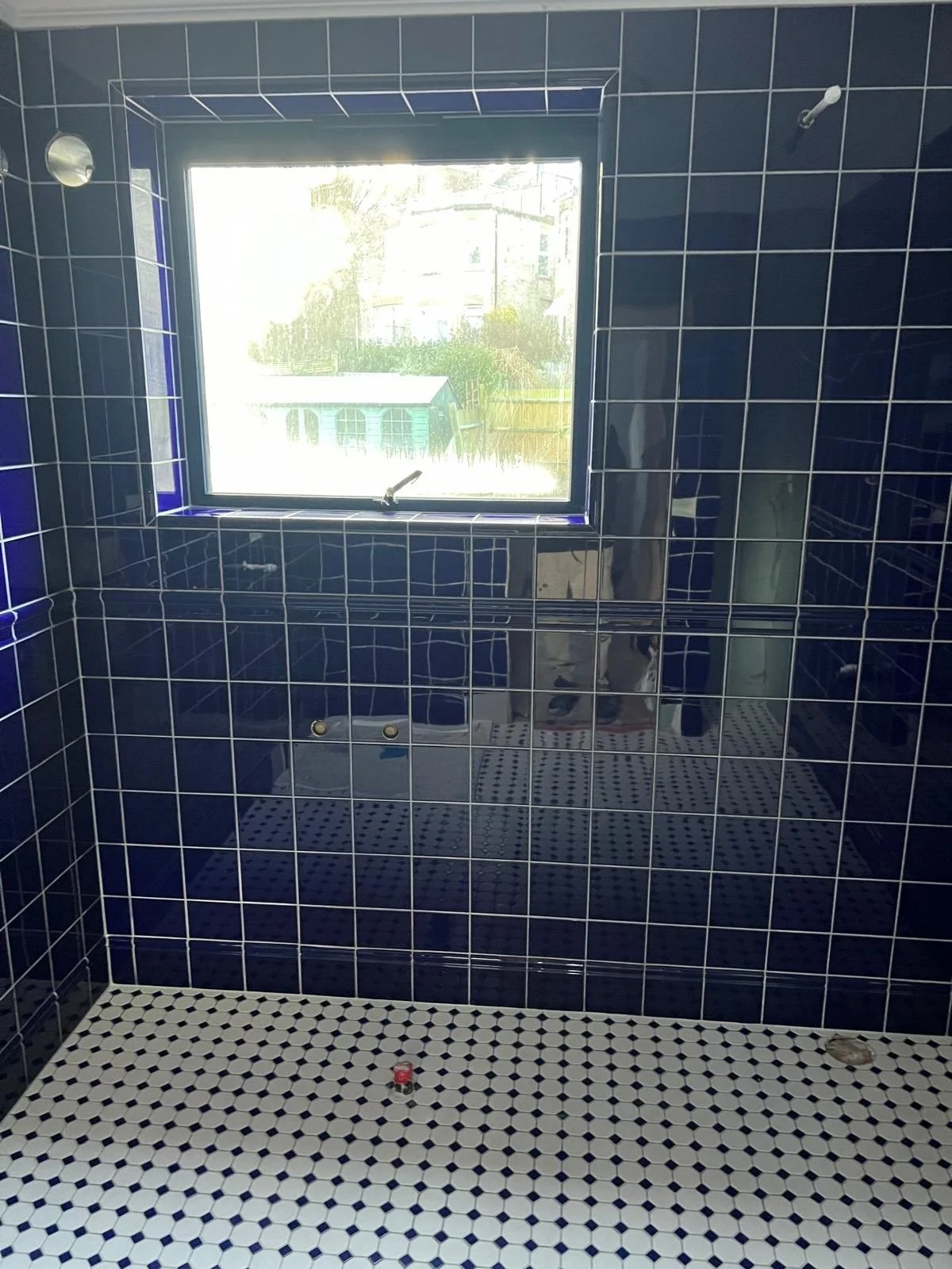 Bathroom with dark blue tiled walls, a window letting in natural light, and black and white small tiled floor. There are two fixtures on the wall, one small round mirror and a hook, with a shadow of a person visible in the reflection.