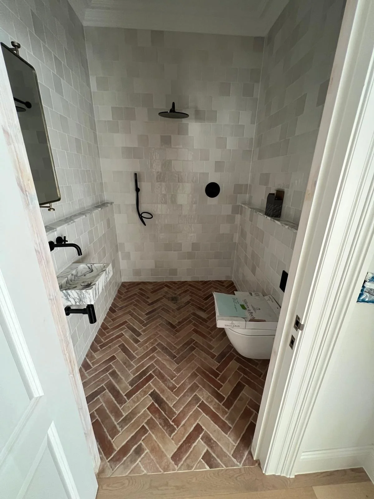 Modern bathroom with herringbone patterned brick floor, white tiled walls, a marble sink with black fixtures, a black showerhead and handheld spray, and a white wall-mounted toilet with a box placed on top. The room appears to be under construction o