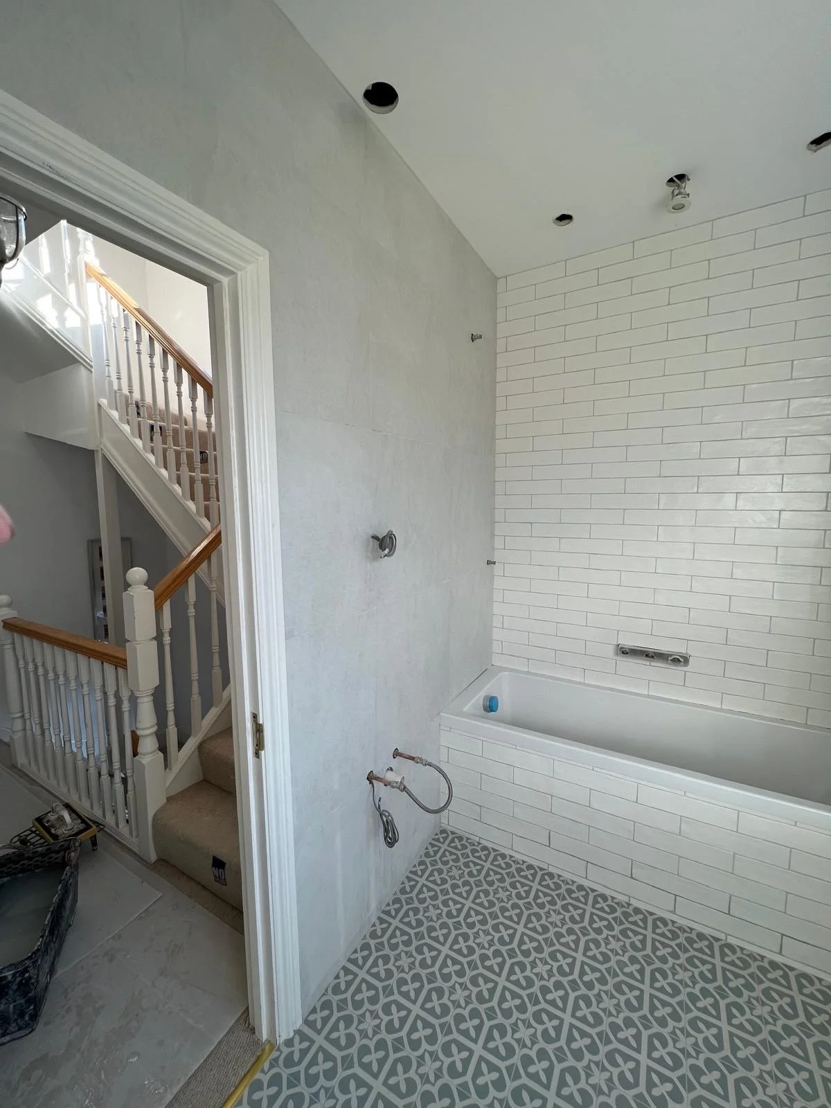 Bathroom under construction with white subway tile on wall, patterned floor tiles, and an empty bathtub. Exposed plumbing and electrical fixtures are visible, and the adjacent hallway shows a staircase with white and wood railings.