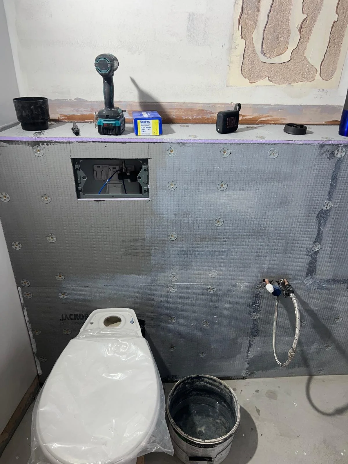 Bathroom under renovation showing a partially installed toilet and plumbing, with tools and materials on the wall behind.