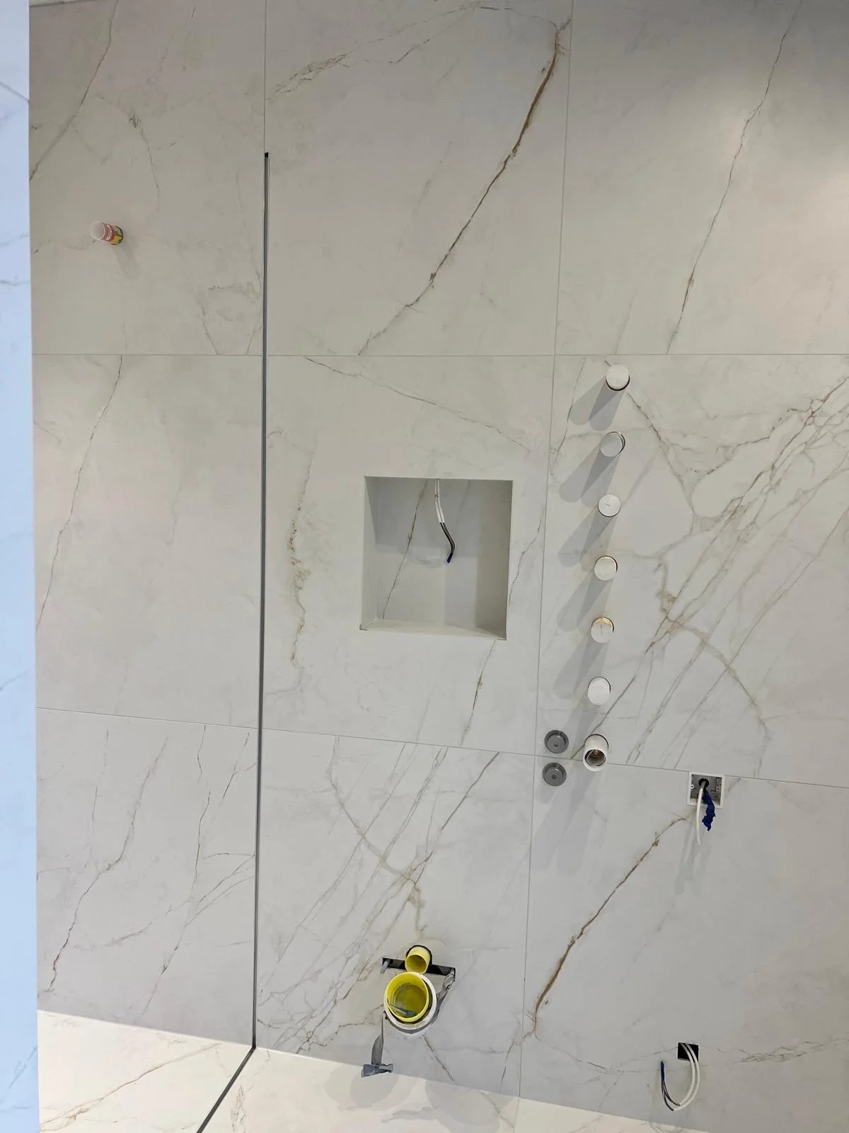 Close-up of a partially installed bathroom wall with marble tiles, showing plumbing fixtures, electrical wiring, and an empty recessed shelf.