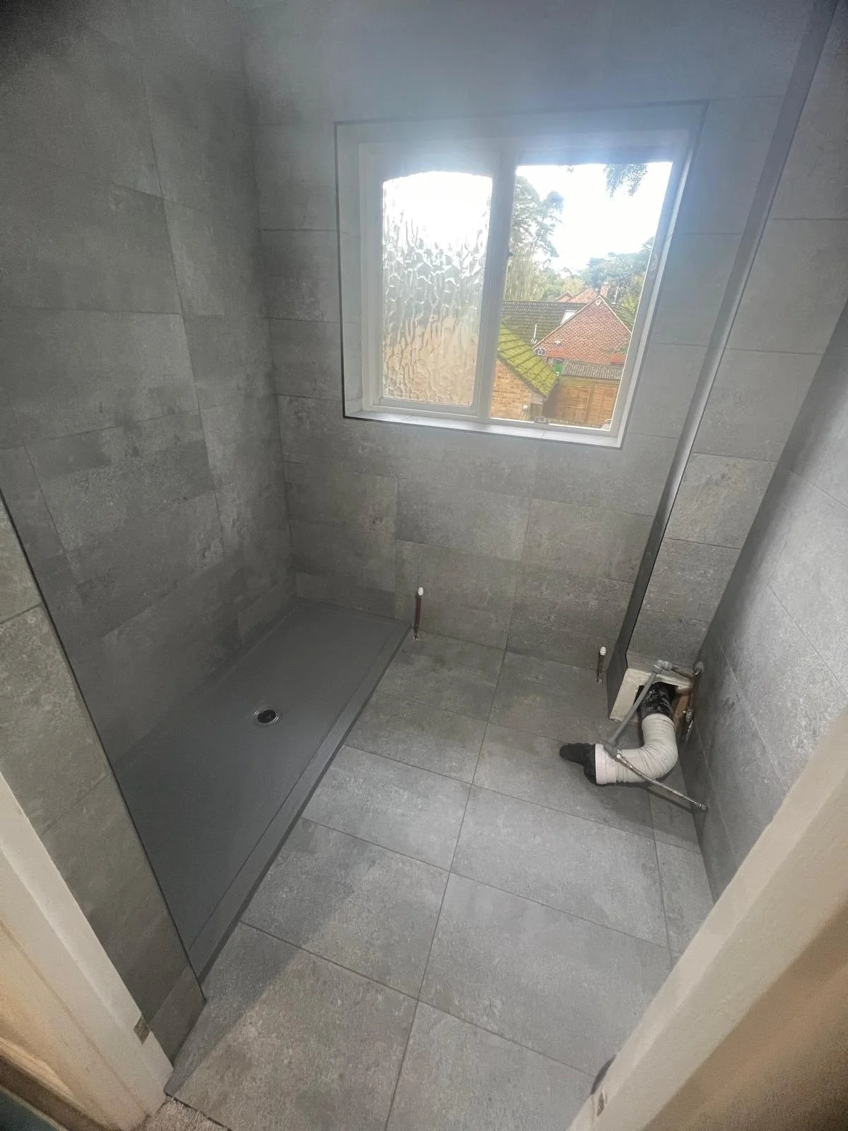 Empty bathroom with gray tiled walls and floor, a window showing outdoor trees and houses, a shower base, and plumbing fixtures.
