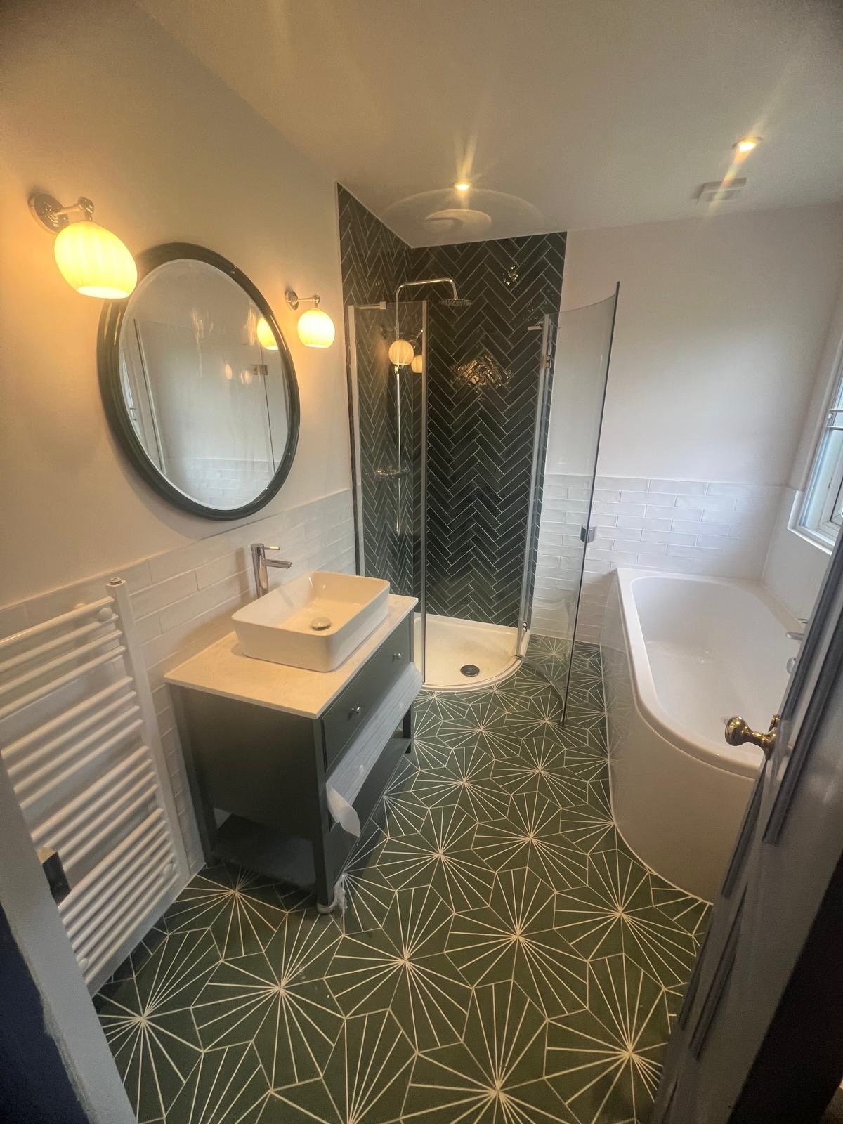 A modern bathroom with geometric patterned floor tiles, a bathtub, a small vanity with a vessel sink, a shower with black tile walls, and a window