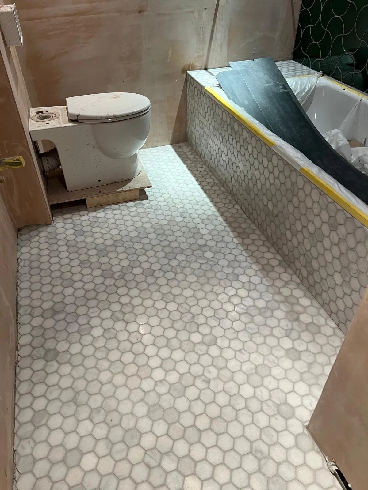A bathroom under renovation with a toilet on a wooden platform, a section of the tub with hexagonal tiles, and construction materials and plastic sheeting around.