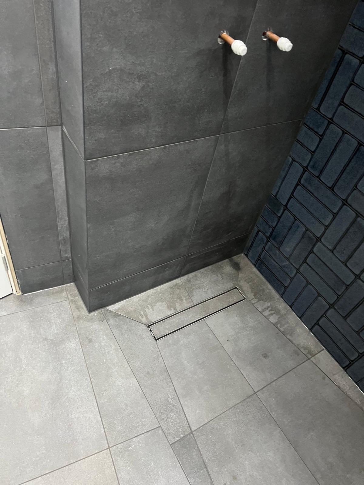 Close-up of a modern indoor space with gray tiled walls and flooring. Two exposed copper pipe fixtures are visible on the wall, and a floor drain with a rectangular cover is on the tiled floor.