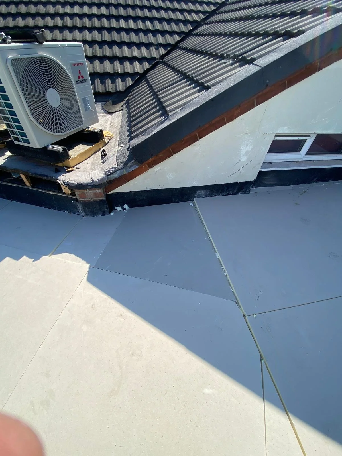 View of a rooftop with an air conditioning unit, roofing tiles, a small window, and a flat rooftop area with tiles and a crack.
