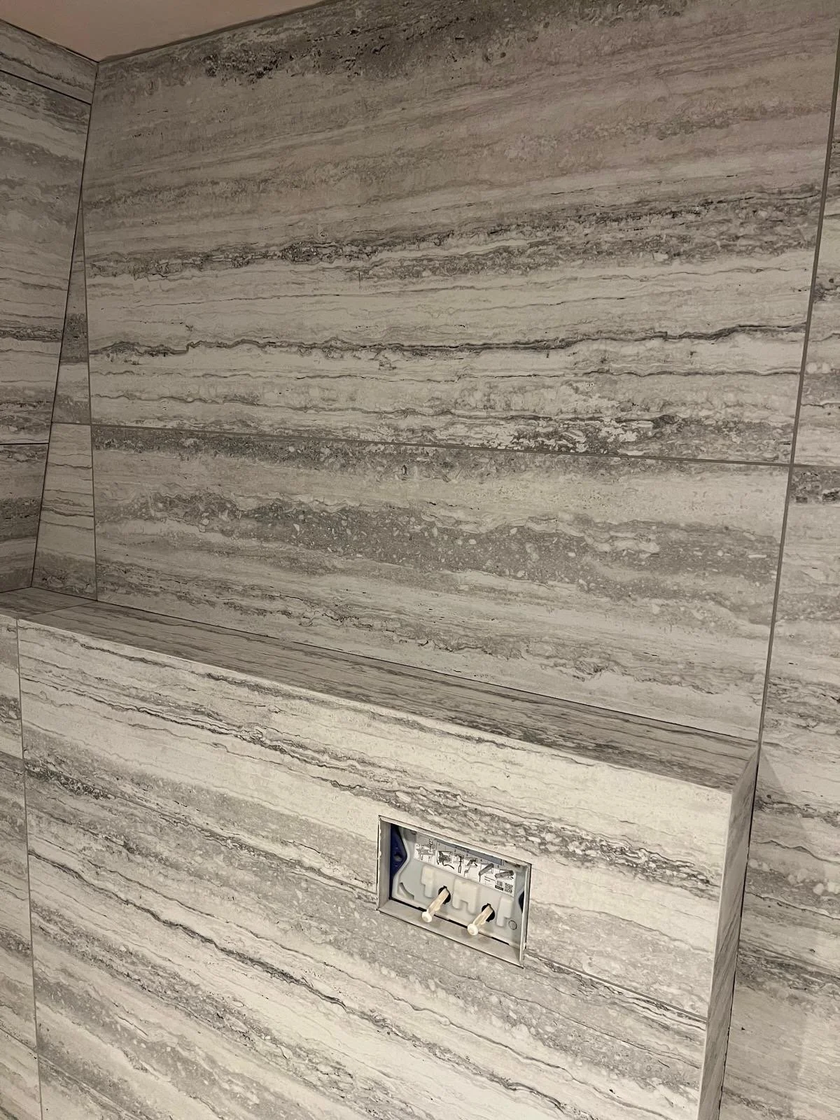 Bathroom wall with beige marble tiles and a recessed electrical outlet.