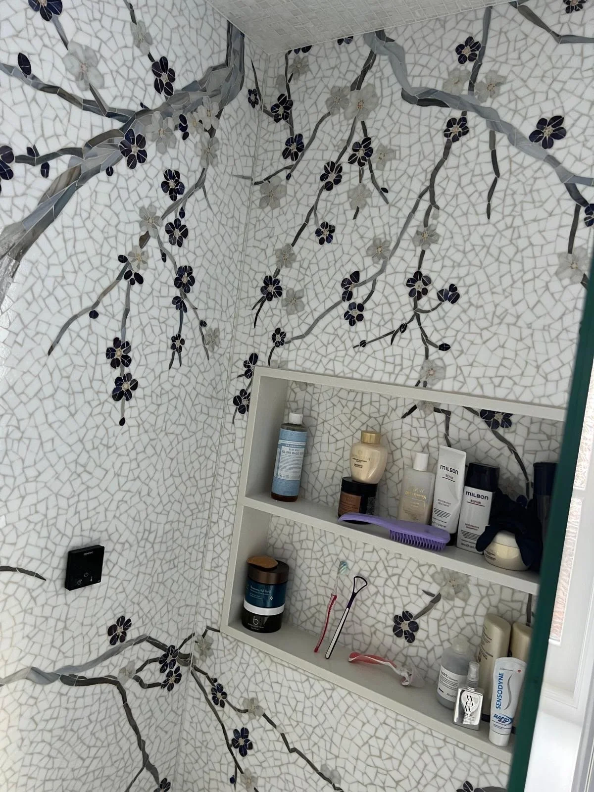 Bathroom corner with floral mosaic tile walls and a white wall-mounted shelf holding various personal care products, toothbrushes, and a mirror.