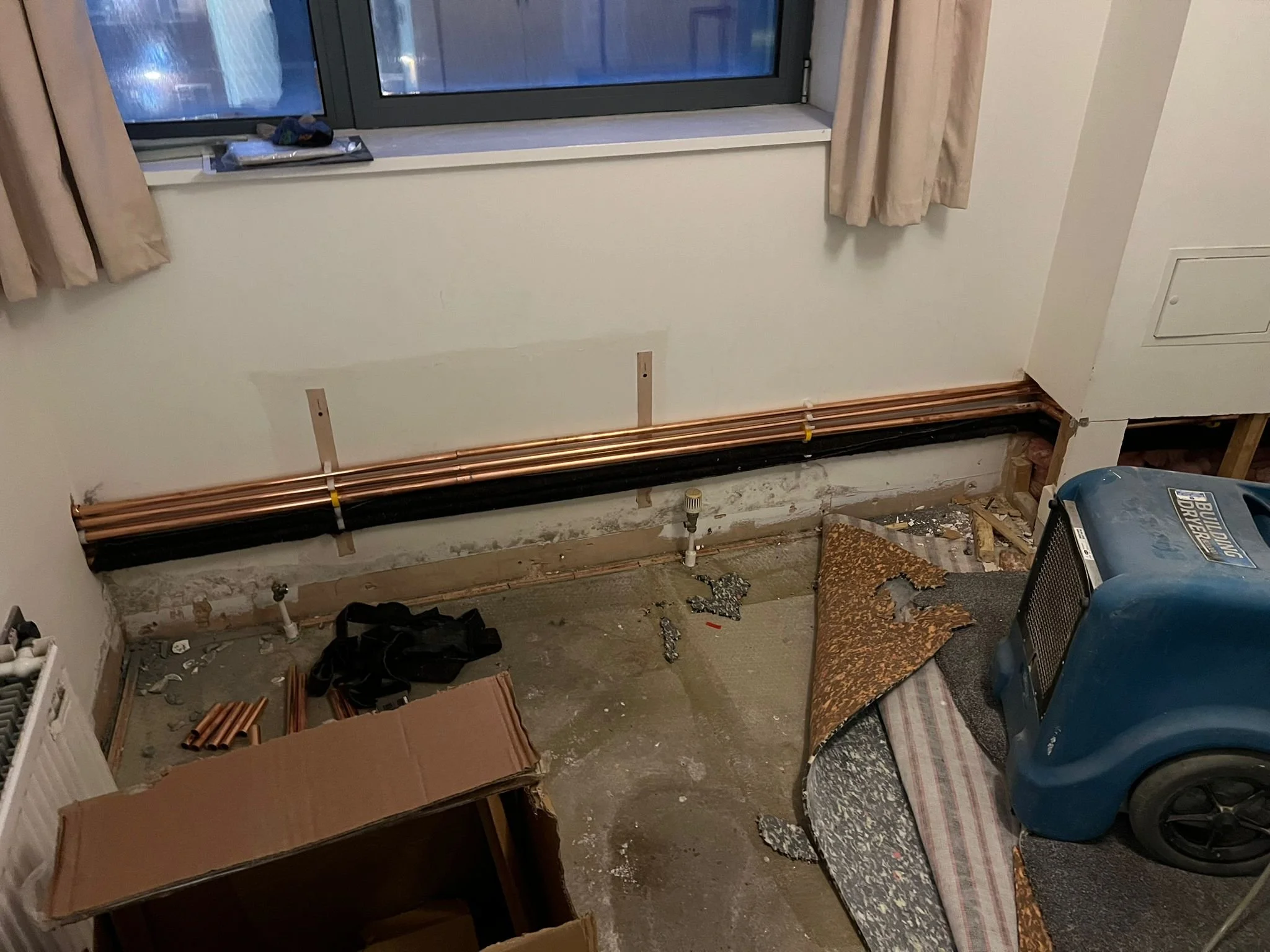 Room under construction or renovation with copper pipes installed along the wall, a window with beige curtains, scattered construction debris, a cardboard box, a blue air compressor, and a small table with a striped cover.