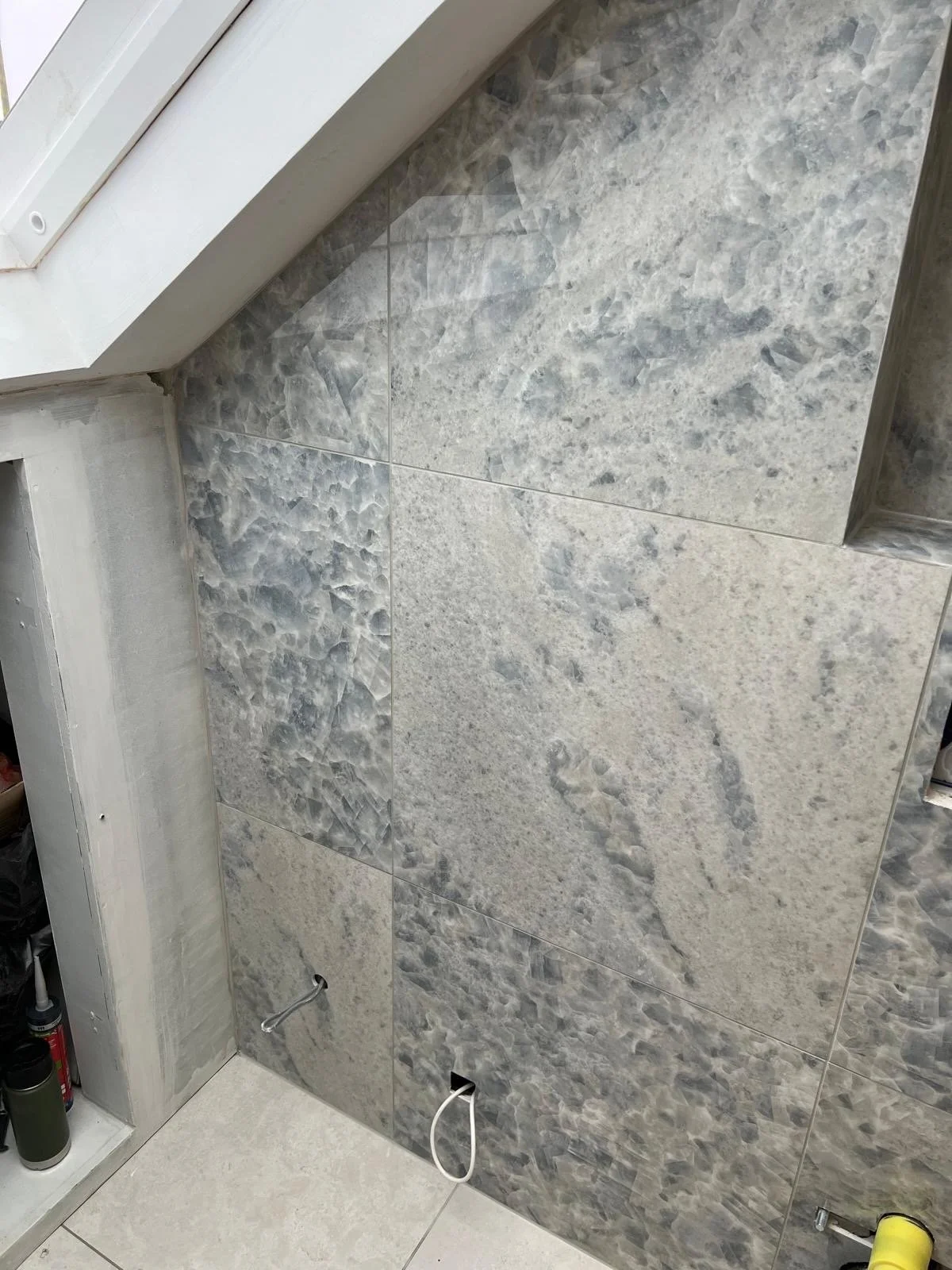 Interior wall with large granite tiles in shades of gray and beige, near a window sill, with visible plumbing fixtures and a small shelf with spray cans.