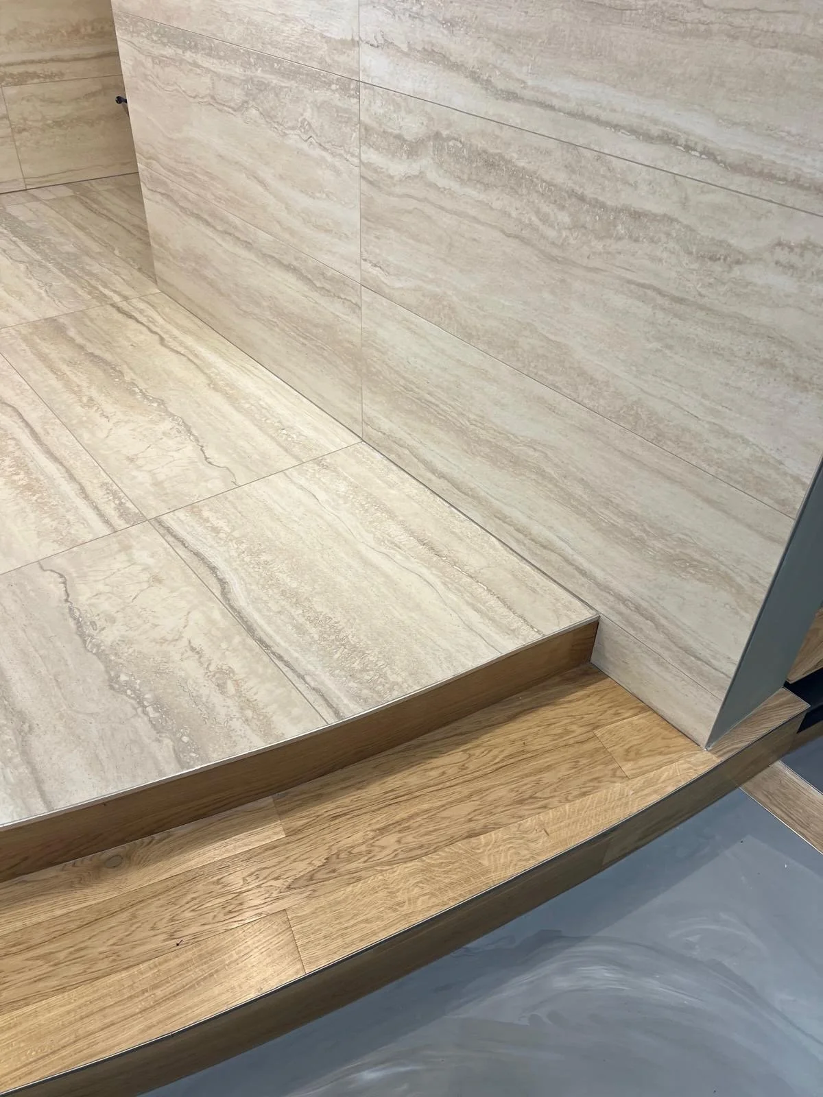 Close-up of a bathroom shower area with beige marble tiles on the wall and floor, bordered by a wooden strip at the base of the wall.