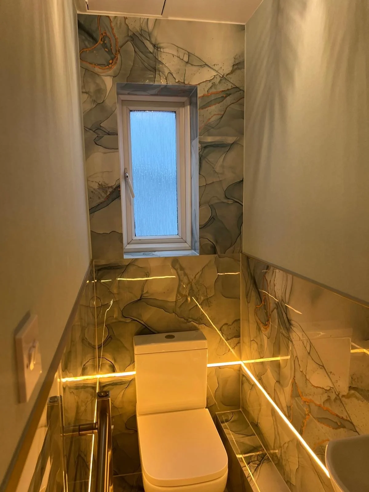 A small, modern bathroom with marble-patterned walls, a frosted window, a white toilet, and LED strip lighting along the edges.