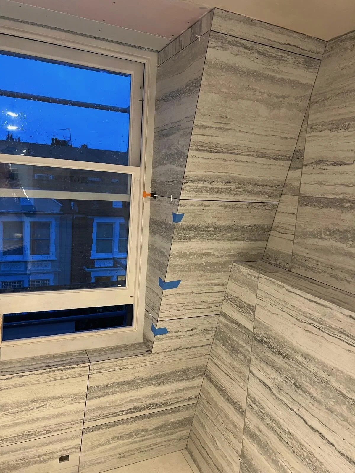 Bathroom with marble tile walls and a window showing neighboring buildings. The tiles have gray and beige veining, and some sections are being installed with blue painter's tape and spacers.