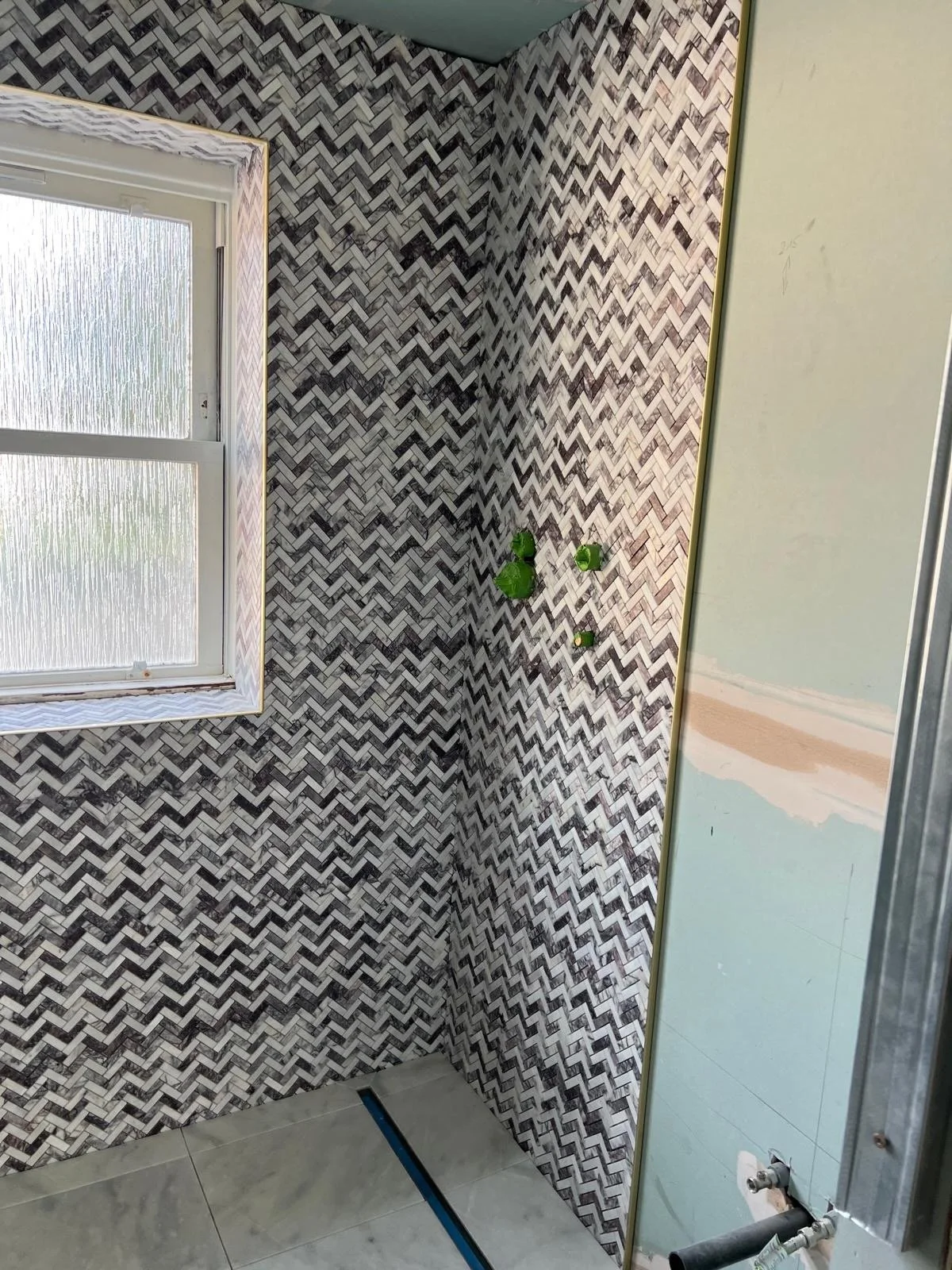 View of a bathroom corner with a window, black and white herringbone tile walls, and plumbing pipes for a sink installation.