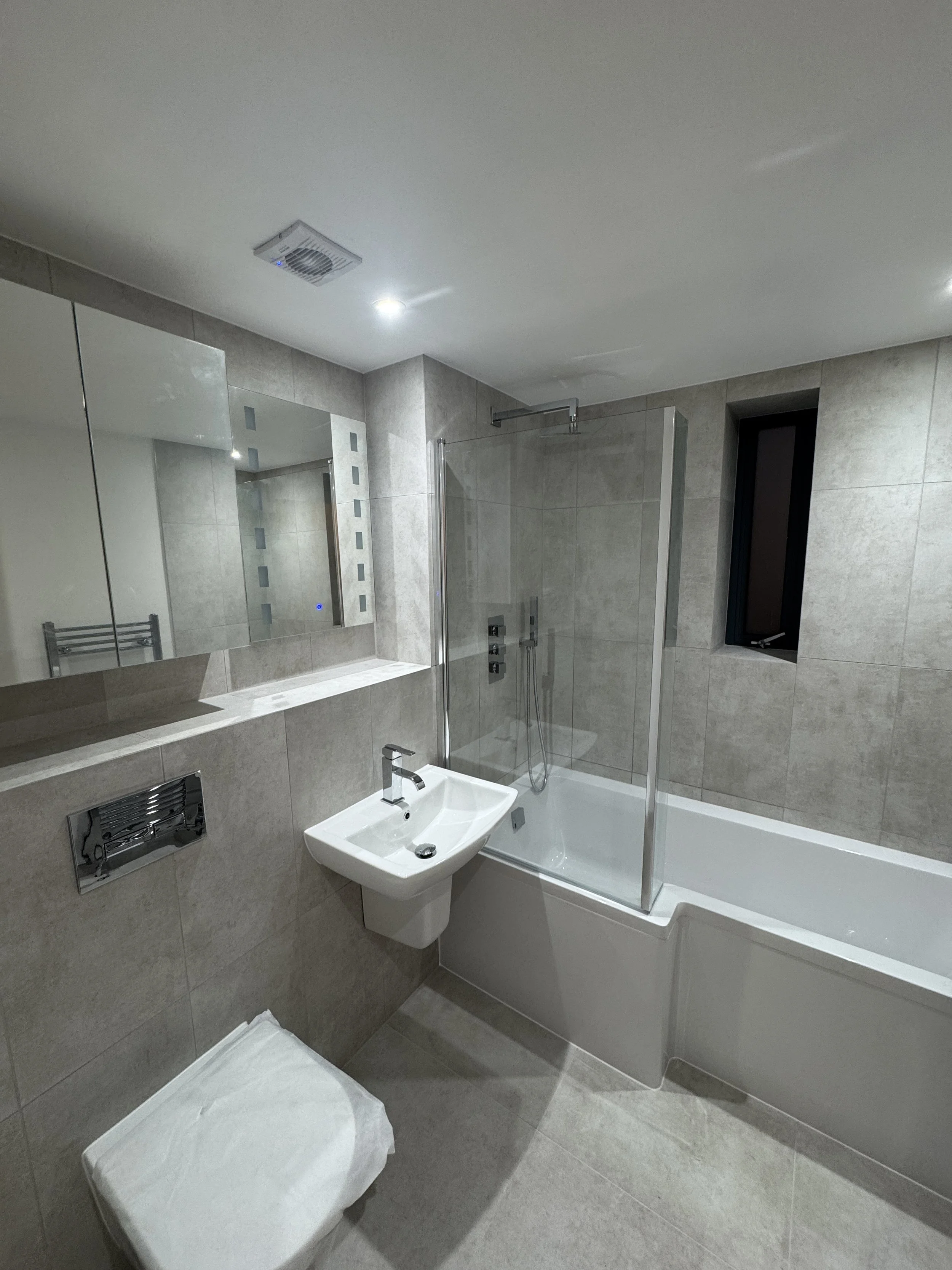 Modern bathroom with gray tiled walls and floor, a white sink, a bathtub with a glass shower enclosure, a large mirror, a small window, and a towel rail.