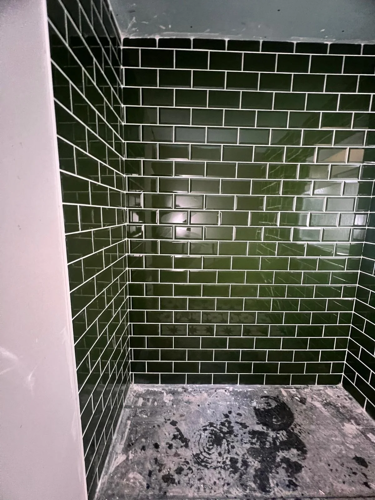 Close-up of a corner with green rectangular subway tiles on the wall, white grout, and a concrete floor with paint splatters.