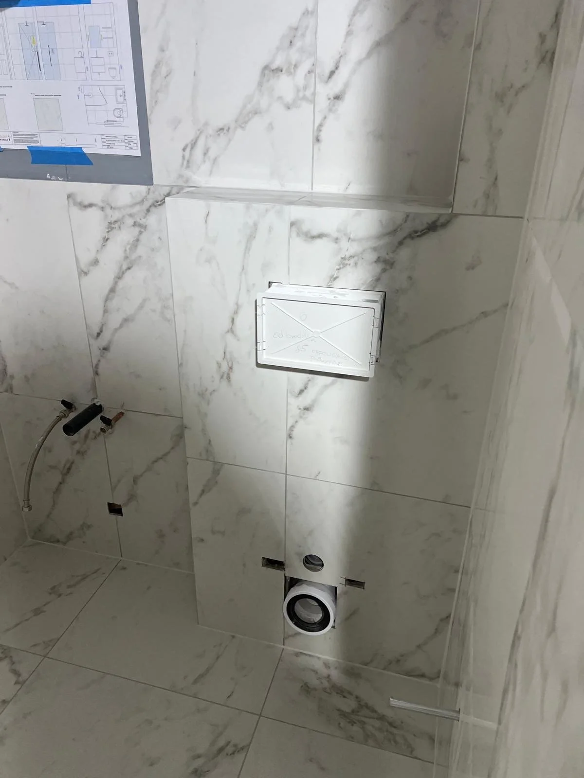 Bathroom with marble tile walls and floor, plumbing pipes and wiring exposed, and an unfinished toilet area with a packaged toilet fixture and handwritten notes on a paper attached to the wall.