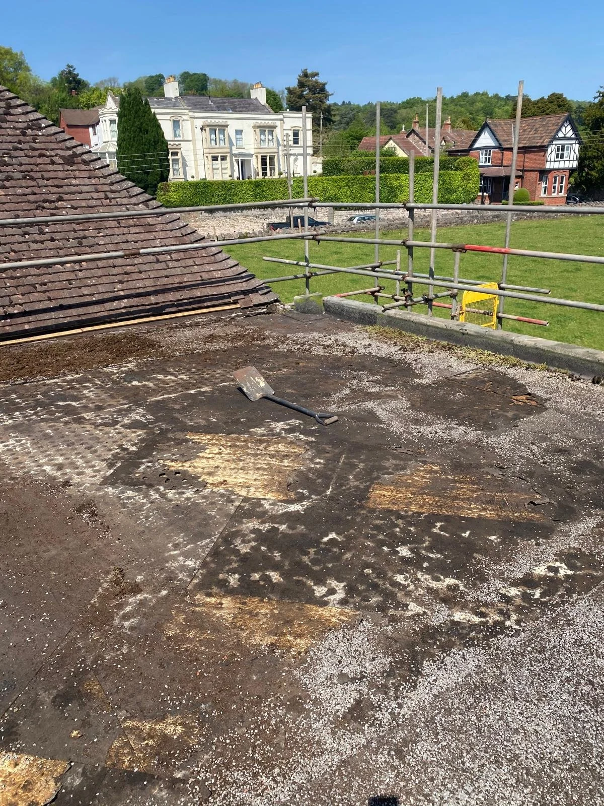 Roof with damaged and worn-out shingles, a trowel lying on the roof surface, and a background of residential houses and green trees under a clear blue sky.