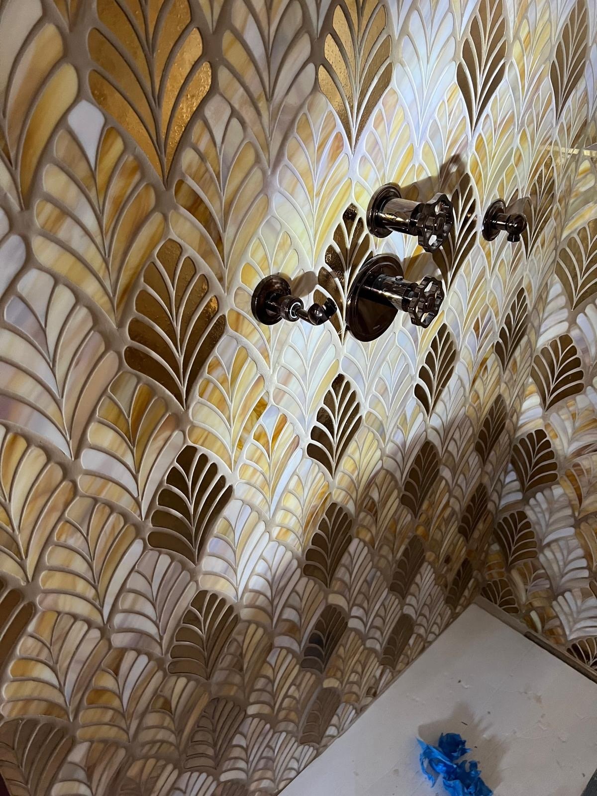 Close-up of a patterned wall with a retro-style fan wall mounted and covered in a white cover. The wall has a decorative tile pattern with various shades of beige, gold, and white in an Art Deco design.