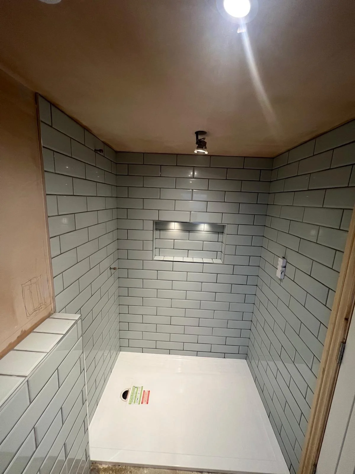 Empty shower stall with gray subway tiles, built-in niche with lighting, and white floor, under construction with visible electrical outlet and plumbing installation.