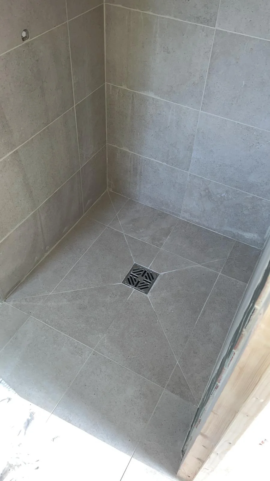 A tiled shower floor with a square metal drain in the center and tan ceramic tiles on the walls and floor.
