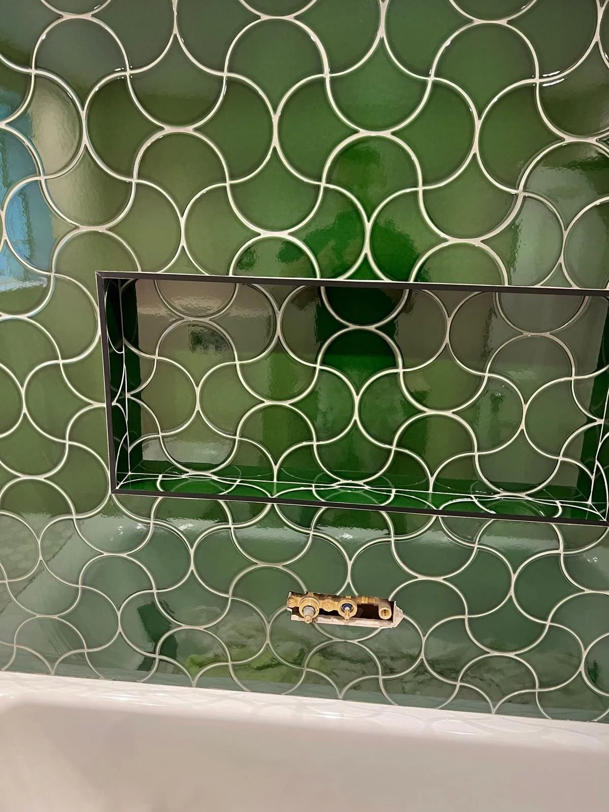 View of a bathroom wall with green patterned tiles and an empty recessed shelf, with an electrical outlet below.
