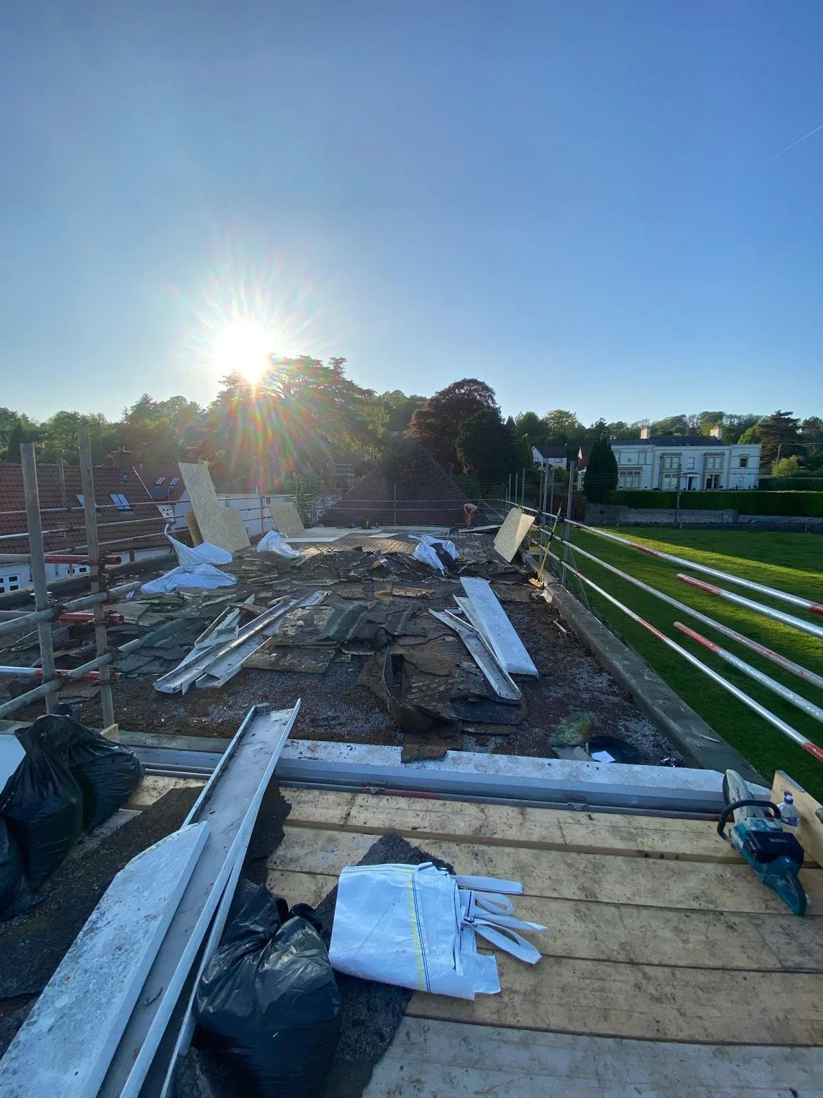 Construction site on a rooftop with building materials, tools, and debris, with a sunny sky and residential houses in the background.