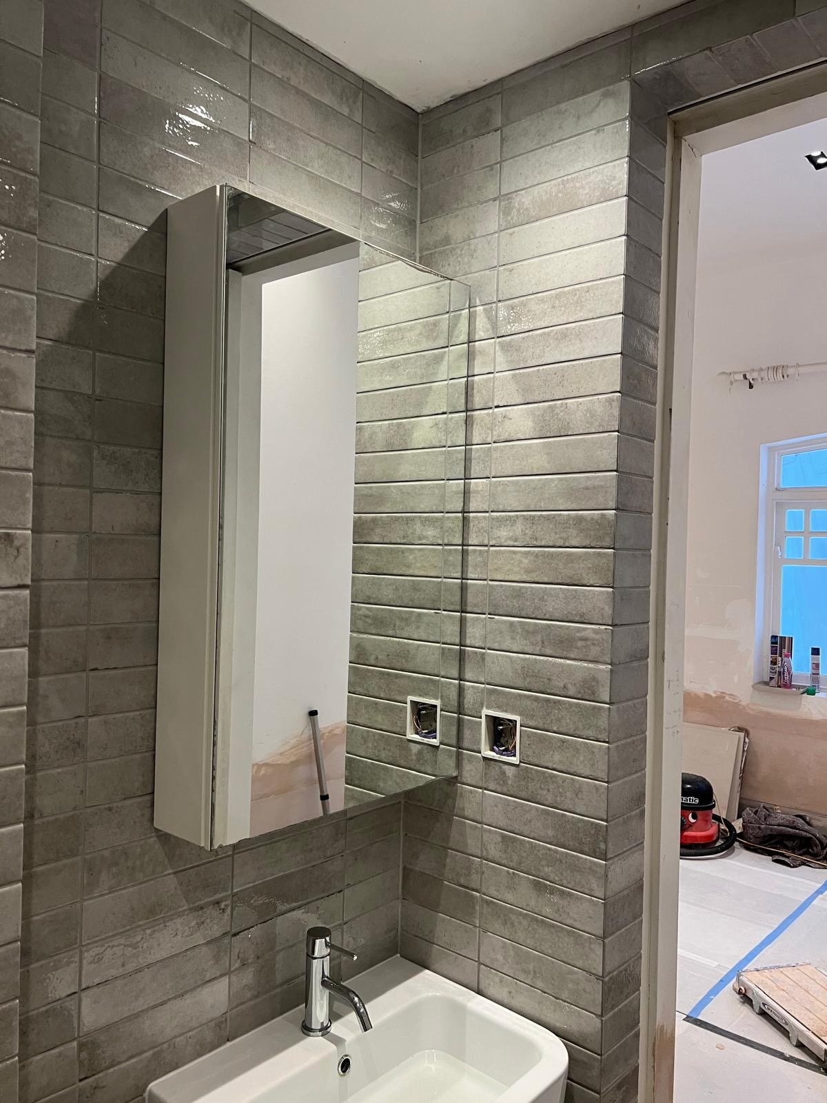A bathroom under renovation with gray tiled walls, a mirror, and a small white sink with a chrome faucet. The area appears to be unfinished with visible electrical boxes and construction tools nearby.