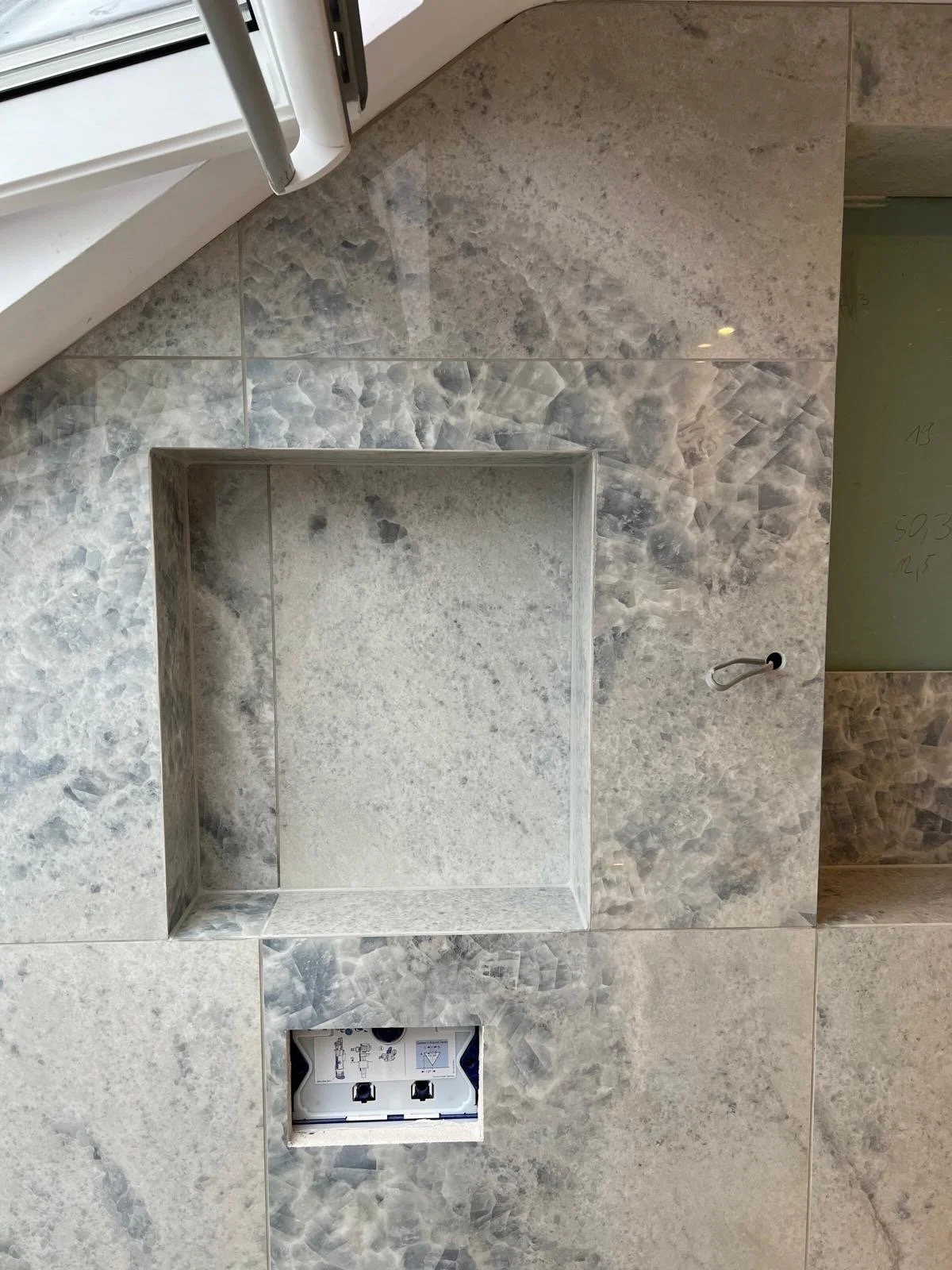 Close-up of a wall with partially installed electrical outlet, surrounded by grey marble tiles, with a recessed niche in the wall and a window at the top left.