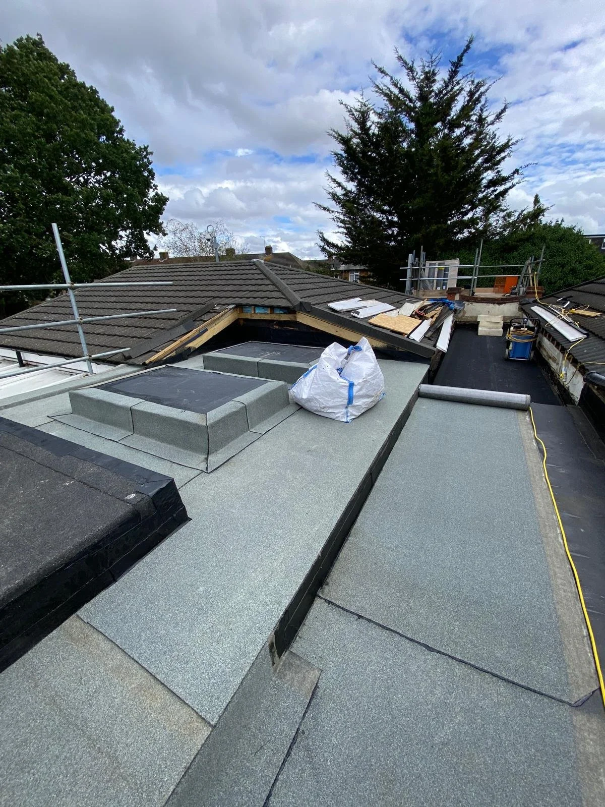 A rooftop under construction showing new roofing materials, tools, and scaffolding, with trees and cloudy sky in the background.