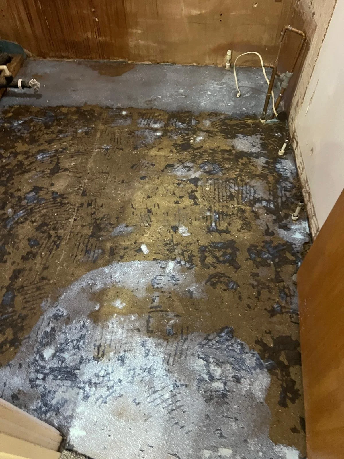 A room with a bare floor that has been stripped of flooring materials, showing an uneven surface with dirt, debris, and footprints. The walls are unfinished, and exposed plumbing pipes are visible in the corner.