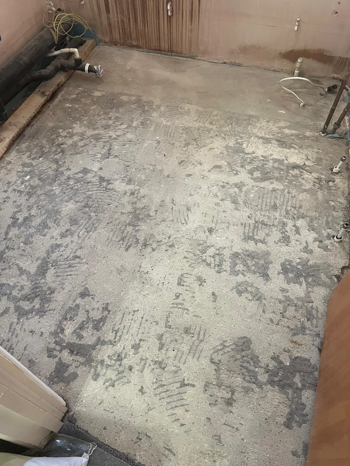 Construction site with a concrete floor covered in foot and tire prints, plumbing pipes on the walls, and electrical wiring hanging.