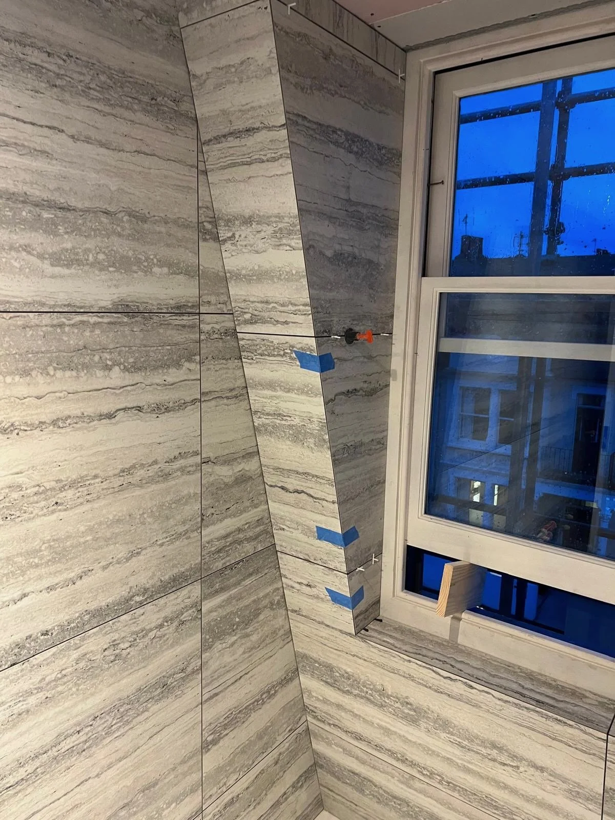 A corner of a room with large gray and white marble or stone tiles on the walls, partially installed. There is a window with a view of a building outside. Blue painter's tape and a black and orange tool are attached to the wall, indicating ongoing co
