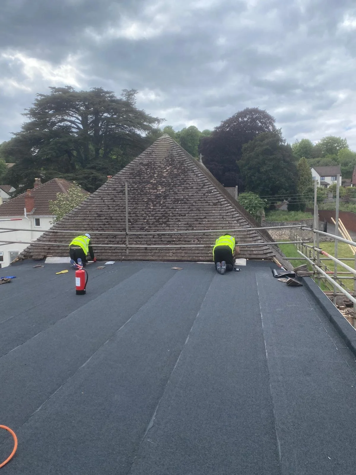 Two workers in bright yellow safety vests kneeling on a rooftop, repairing or inspecting the roof under cloudy skies, with trees and residential buildings in the background.