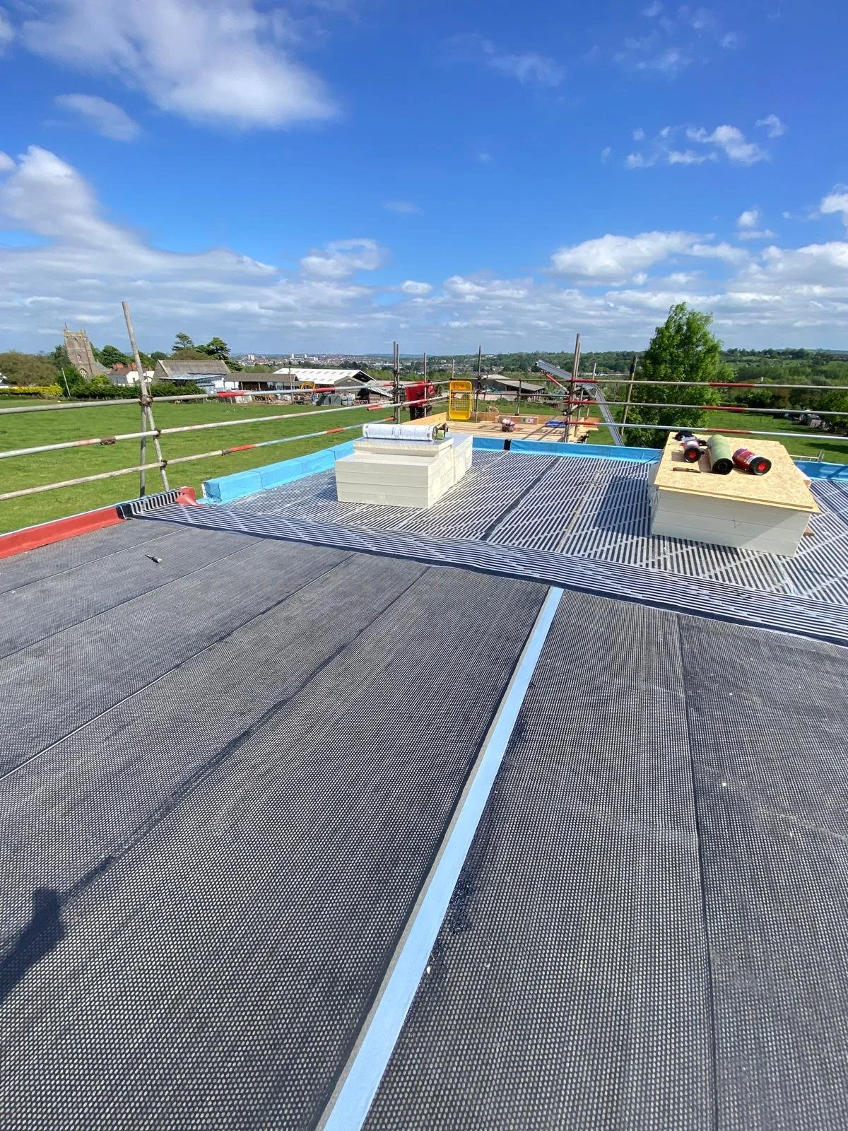 Construction site on a roof with insulation and weatherproofing materials, scaffolding, and safety railings, overlooking a rural area with houses and trees under a partly cloudy sky.