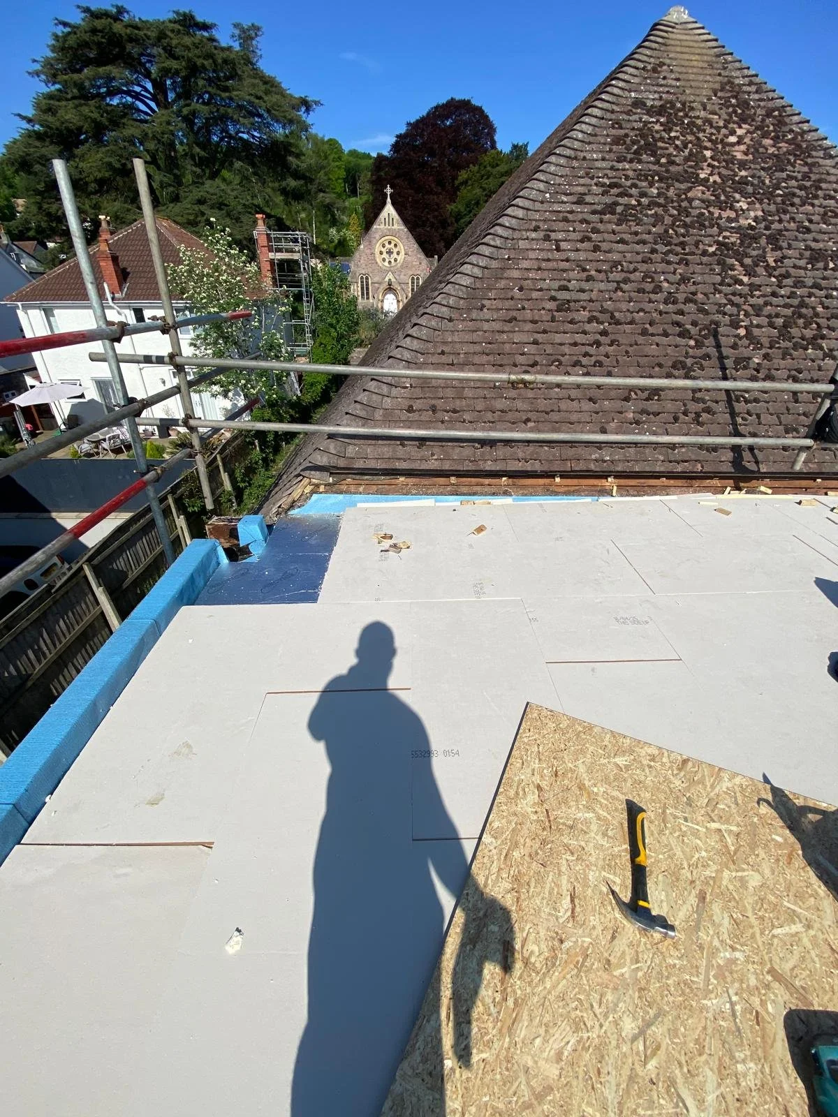 Roof construction site with scaffolding, with an existing sloped tile roof and a new flat roof being installed. There are trees, a church, and neighboring houses in the background.