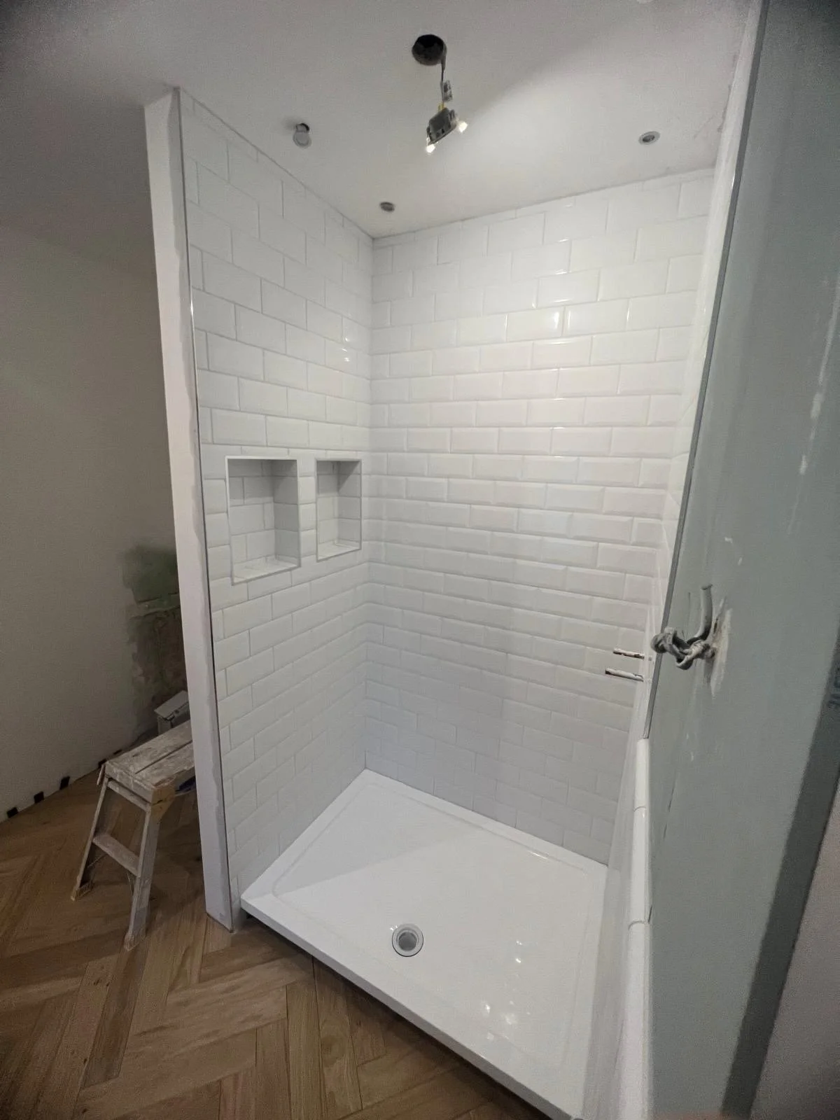 Newly installed white tiled shower with built-in niche shelves, a white shower base, and partial plumbing fixtures, in a bathroom under construction.