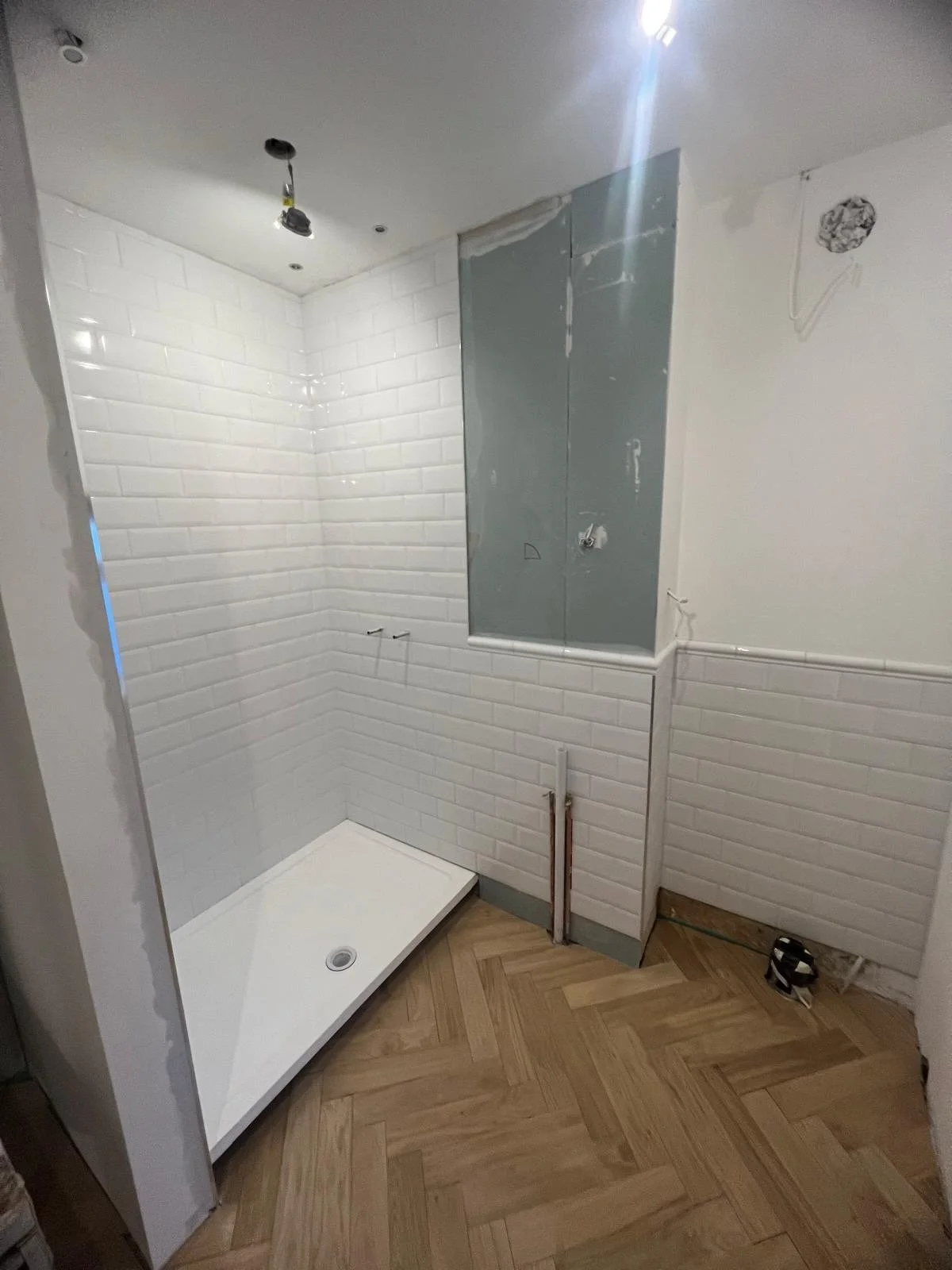 Unfinished bathroom with white subway tile walls, a shower base installed, and plumbing visible on the wall. The ceiling has recessed lighting, and the room is under construction with an electrical outlet and wiring exposed.