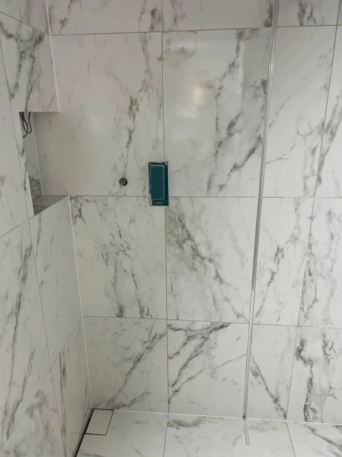 A bathroom with marble-patterned tiles and a built-in shelf. There is an empty blue soap dish on the tile wall, and a visible pipe or drain hole.