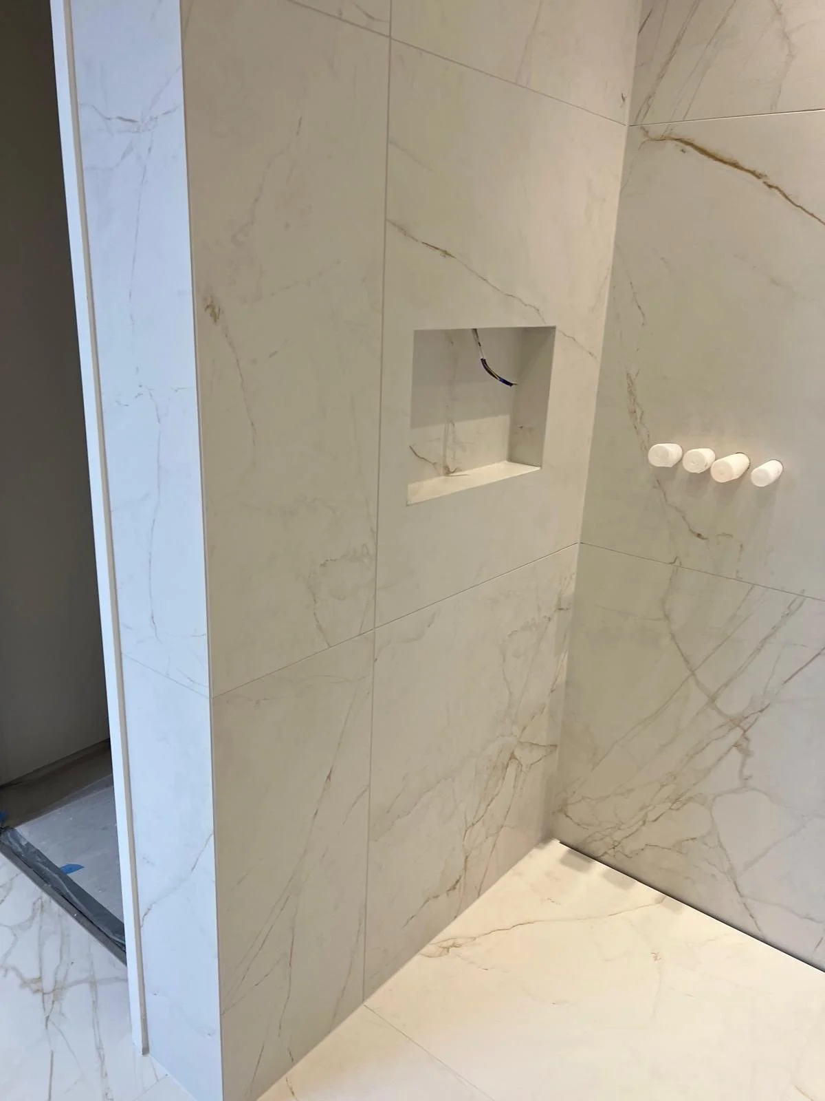 Newly tiled shower with marble walls, a built-in niche, and three white towel hooks.