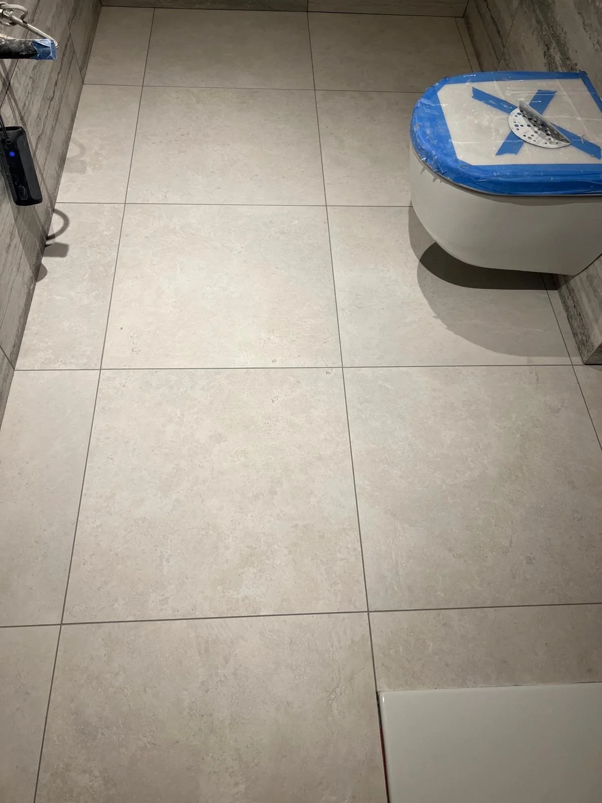 A bathroom with large light beige tiles on the floor, a wall-mounted toilet with a blue cover and tape, and some construction tools and materials nearby.