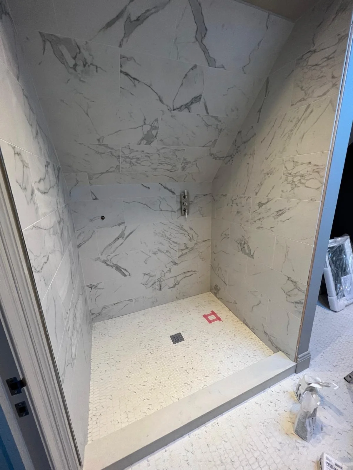 A shower space with white marble tiles on the walls and a textured white tiled floor, with a drain in the center and an unfinished shower fixture on the wall.
