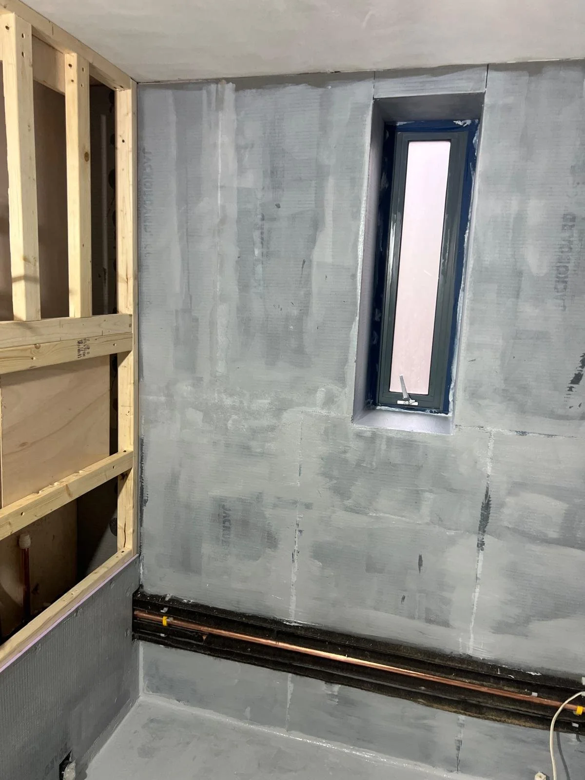 Interior of a room under construction with unfinished drywall, a narrow vertical window, and exposed plumbing pipes along the base wall.