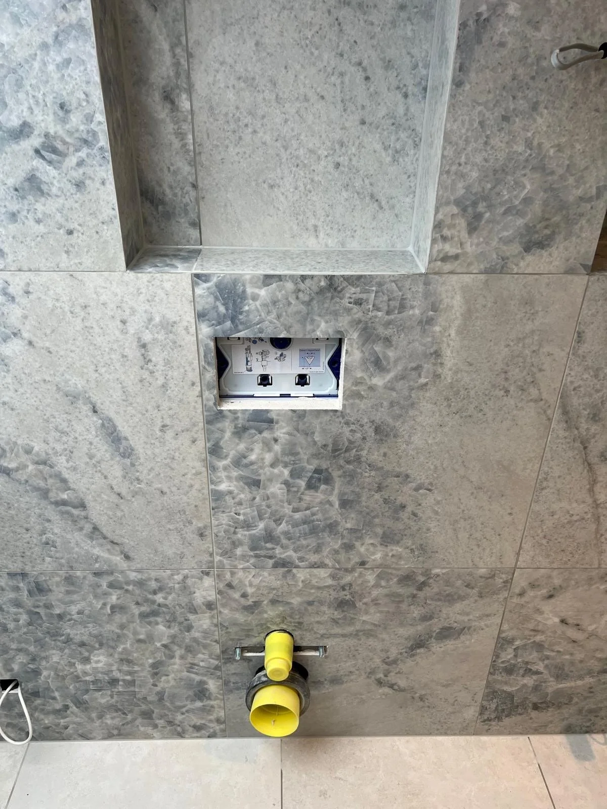 A partially tiled bathroom wall with an electrical box and plumbing fixtures, including a yellow pipe and a drain pipe, on the floor.