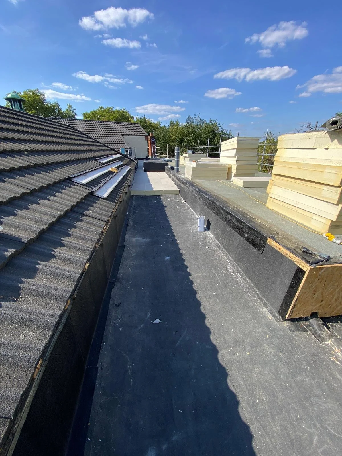 Rooftop under construction with stacks of insulation foam boards, roofing material, and tools, under a blue sky with clouds.