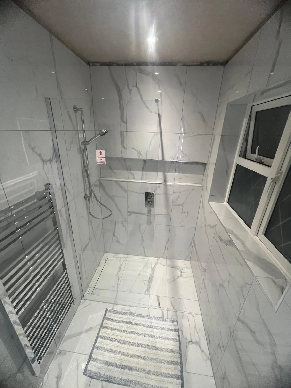 A modern bathroom with white marble walls and a window with frosted glass. It features a handheld showerhead, a towel warmer, and a small striped rug on the floor.