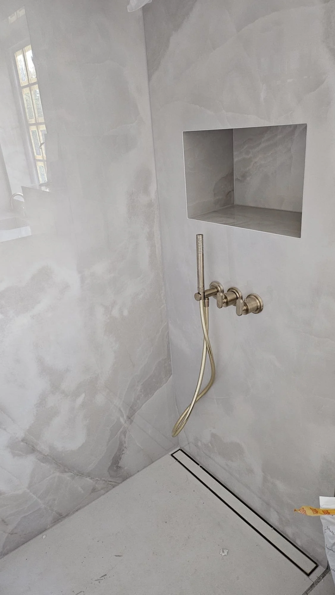 A modern bathroom shower with marble walls, a built-in marble shelf, a silver shower fixture with a handheld showerhead, and a floor drain.