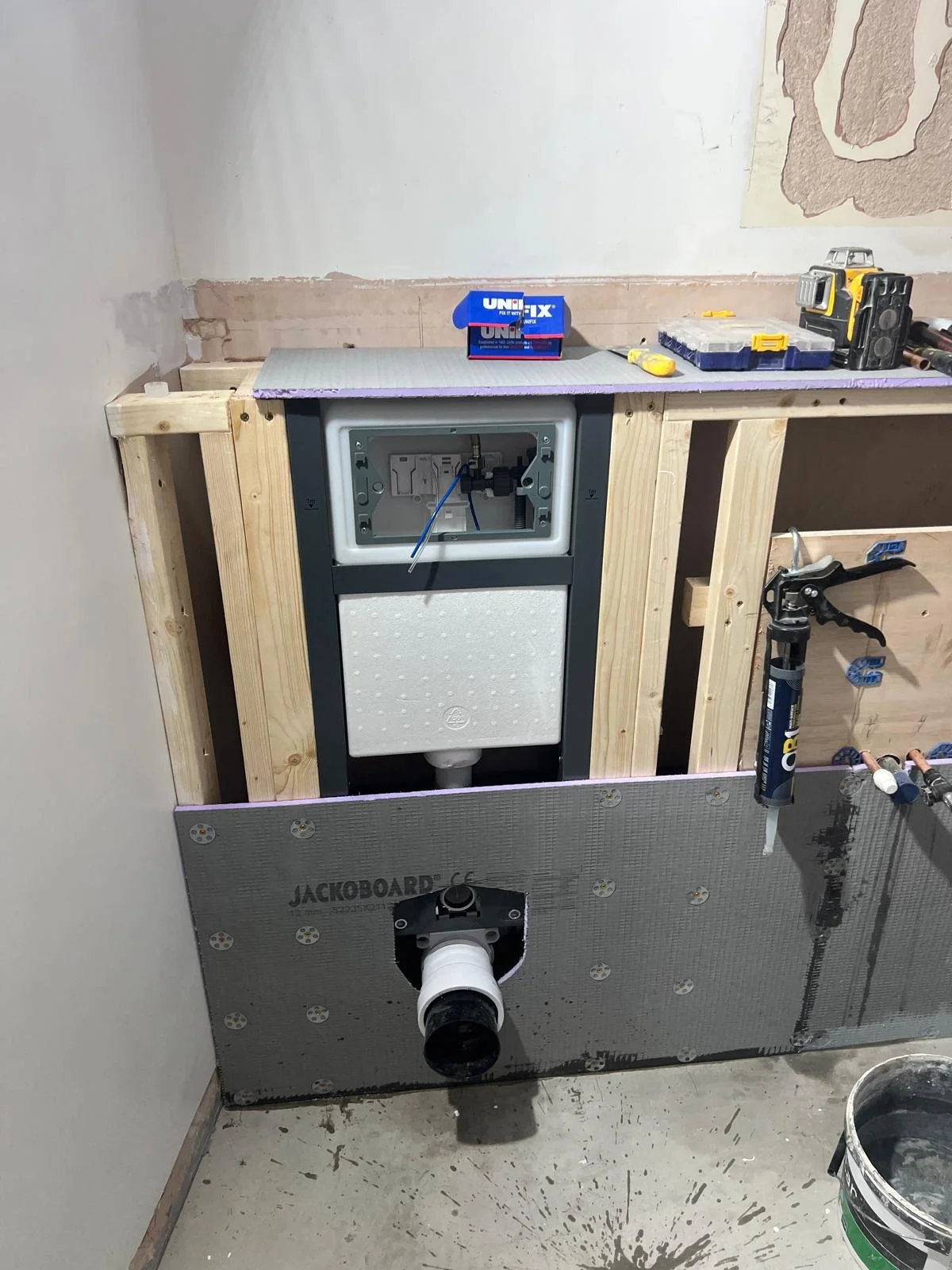 Interior of a bathroom under construction showing a toilet frame and plumbing, with tools and materials on a wooden ledge.