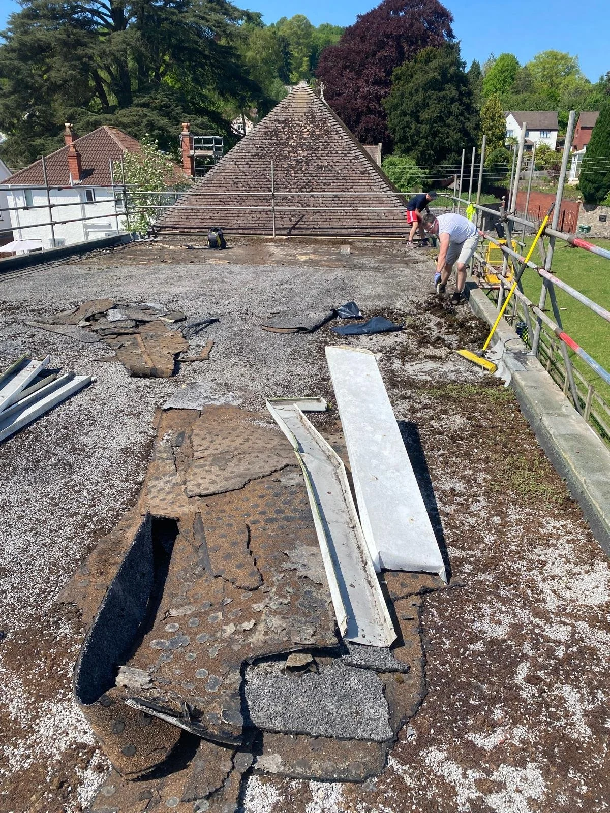 Roof under renovation with debris, tools, and workers removing old roofing materials in a residential area with trees and neighboring houses in the background.